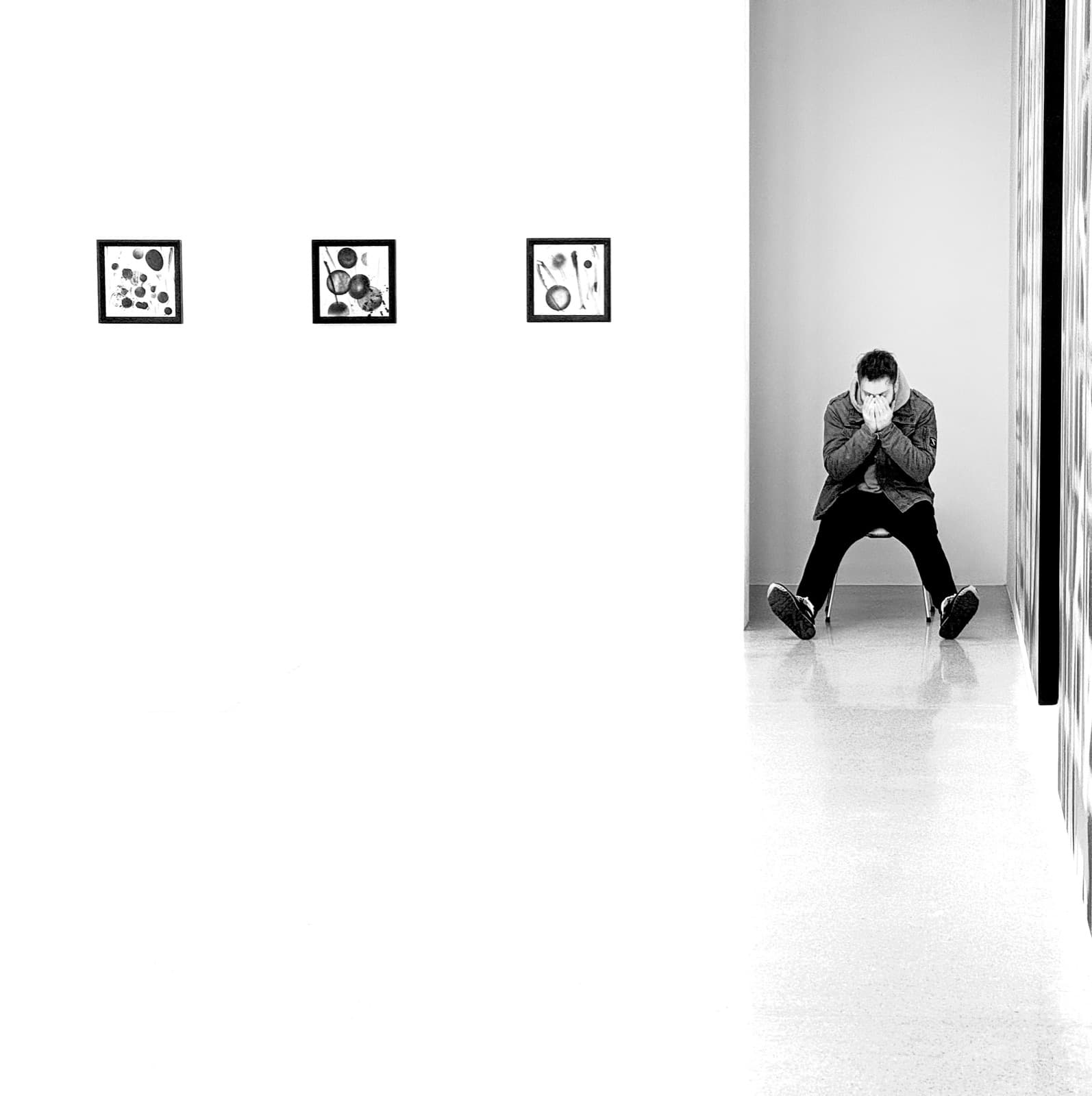 A figure sitting alone against a white gallery wall, head down and hands over the face, beside three small framed works.
